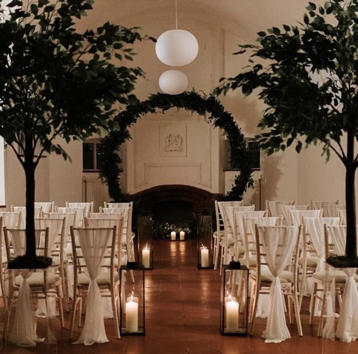 The hall set for a wedding ceremony, with a foliage arch, candle lanterns and chairs either side of the aisle