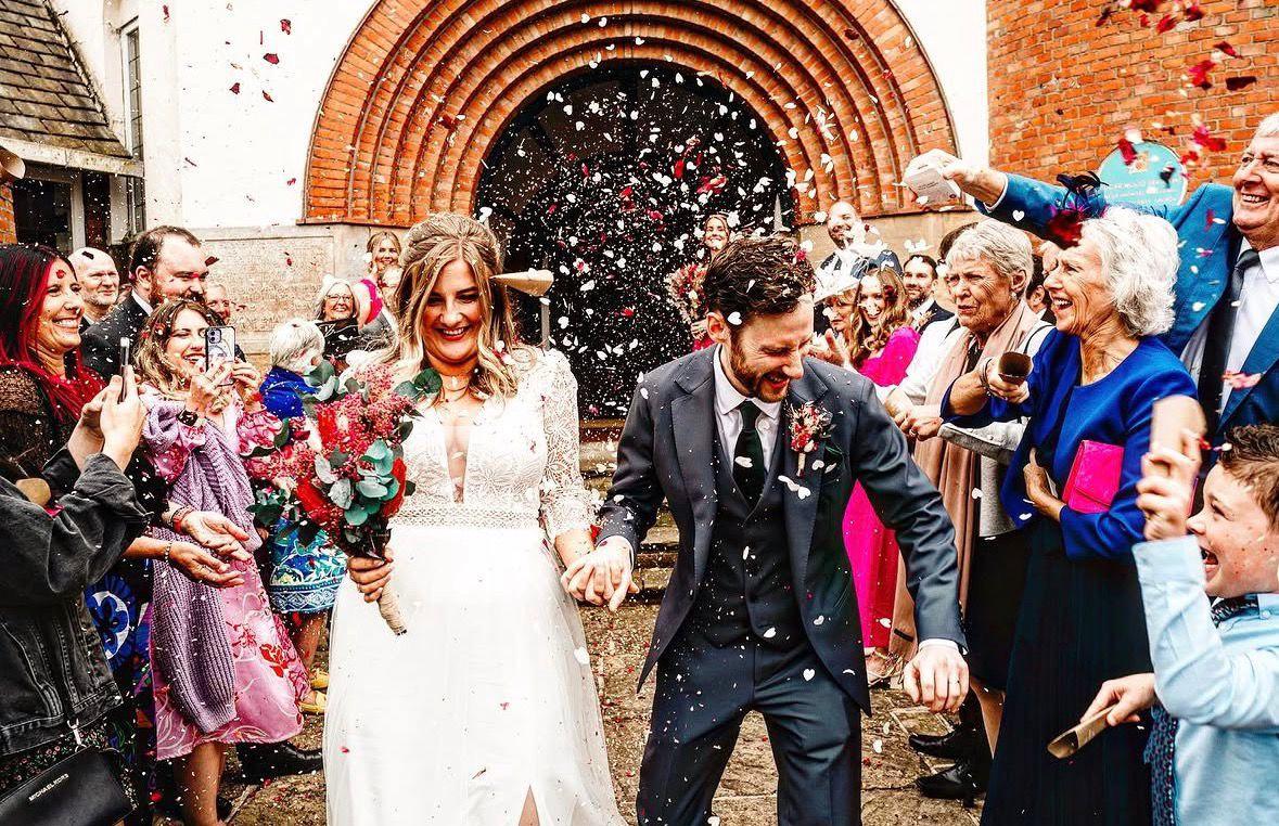 A bride and groom walking hand in hand through a shower of confetti, cheered on by guests on both sides, beneath the brick arch of the entrance