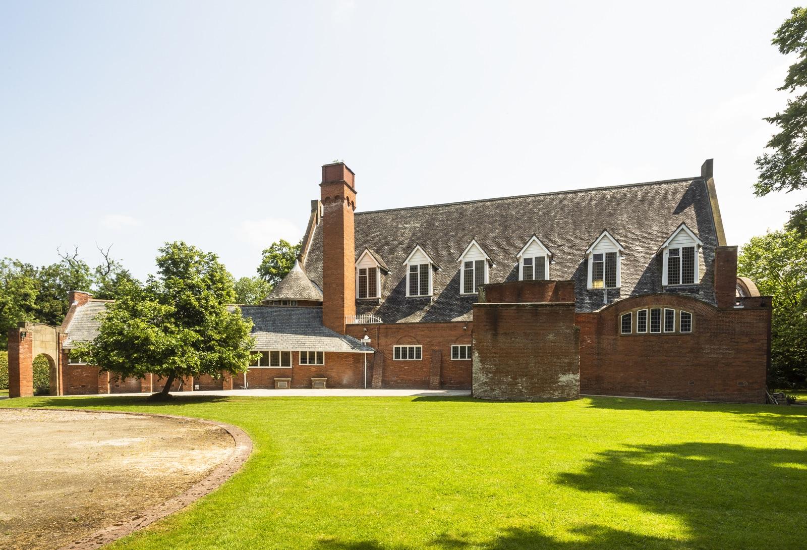 The garden front seen across a wide lawn, with a long slate roof, dormer windows and a tall brick chimney above red brick walls