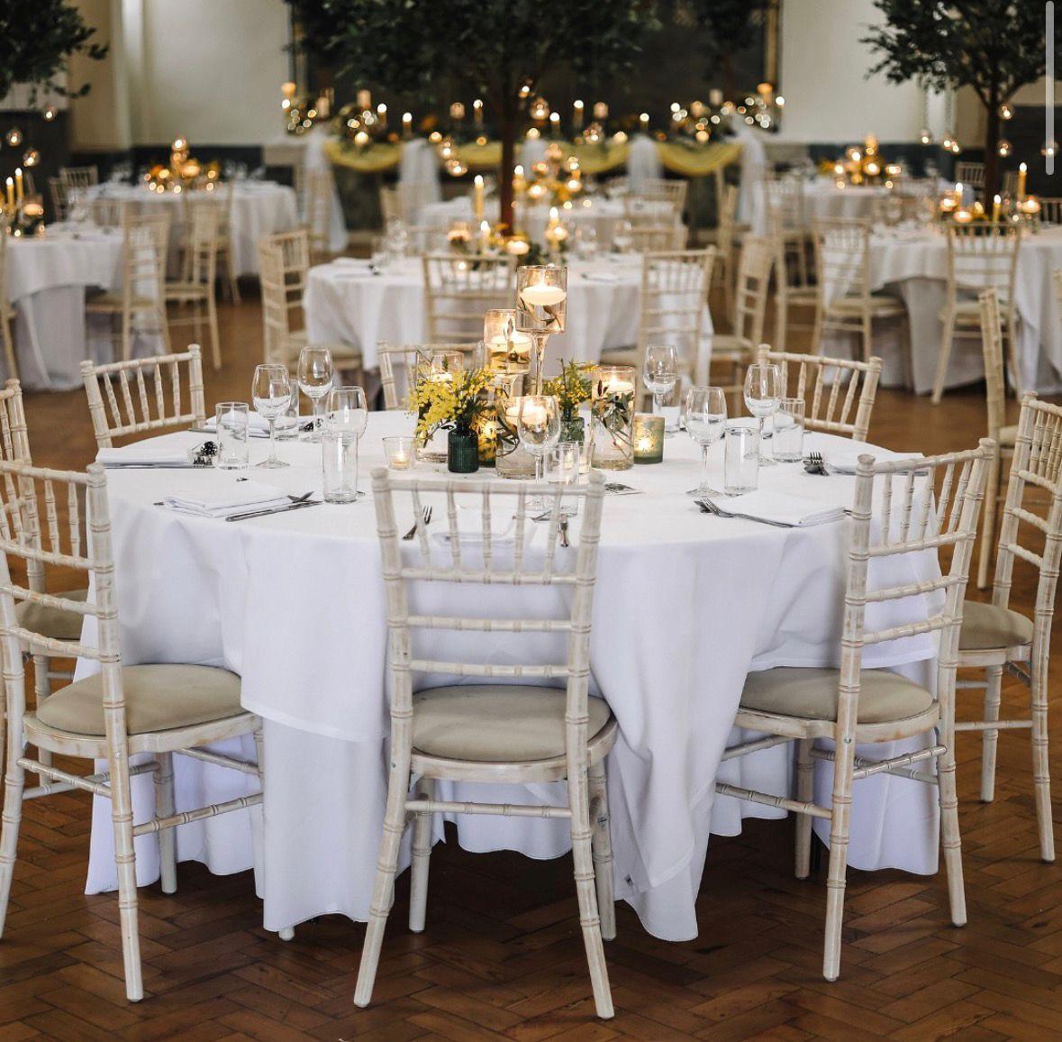 Round tables laid with white linen, glassware, candles and yellow flowers, ringed by white chiavari chairs, with more candlelit tables behind